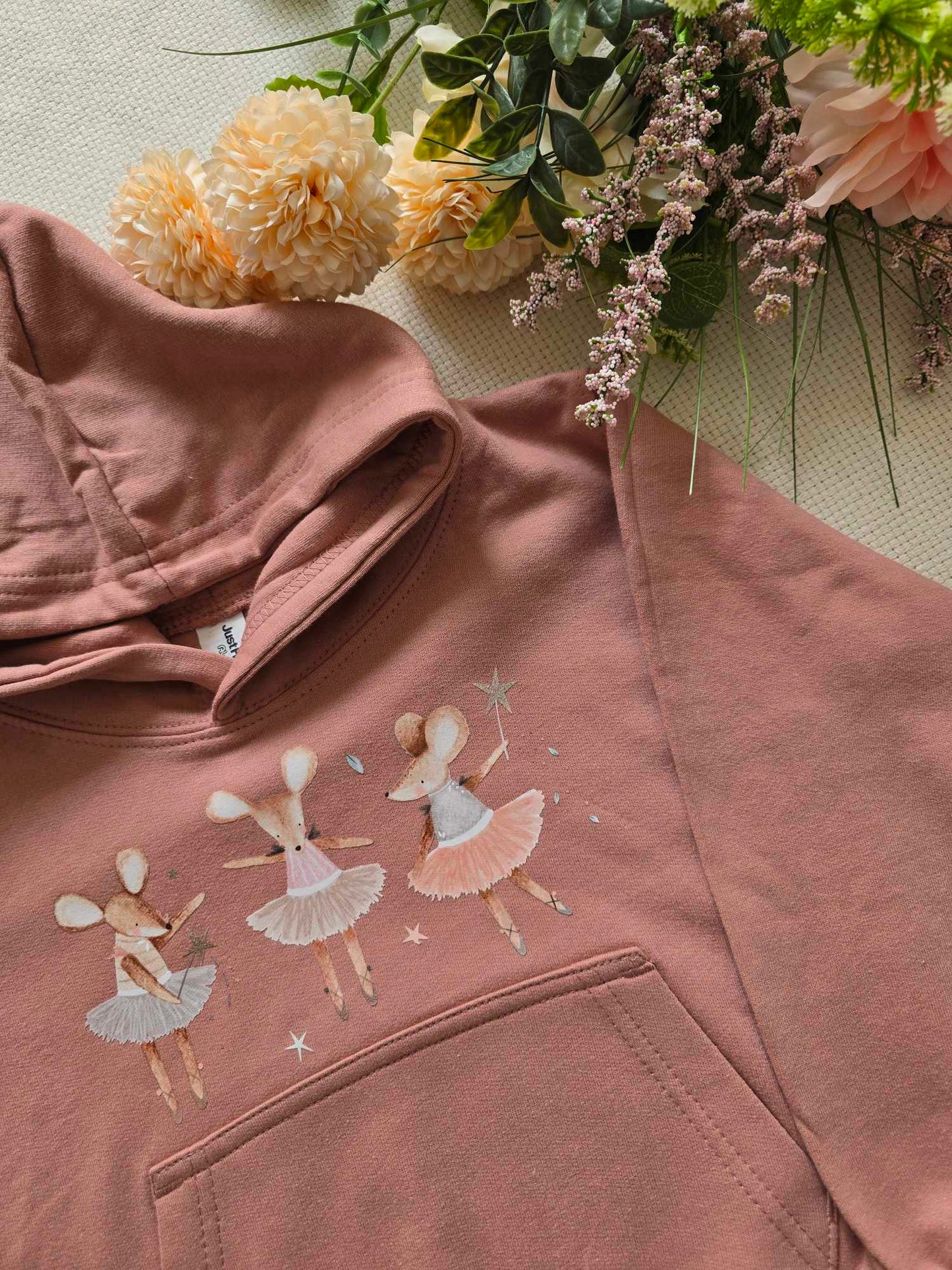 A pink sweatshirt with an embroidered design of ballet dancers and mice, presented alongside flowers.
