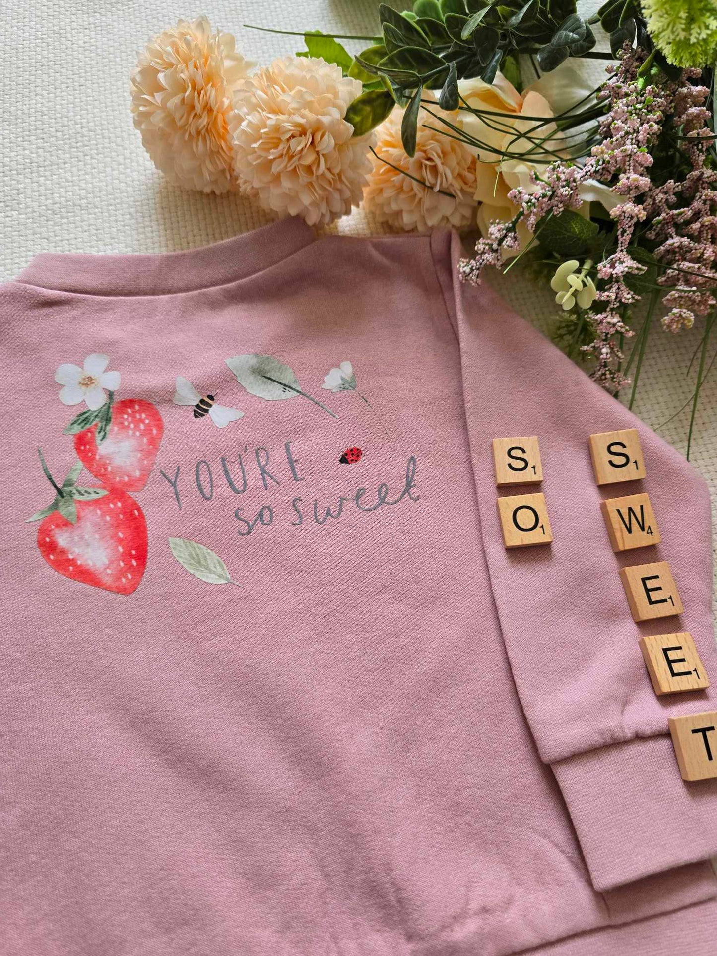 A pink sweatshirt with a text and strawberry graphic design, displayed alongside floral arrangements and strawberries.