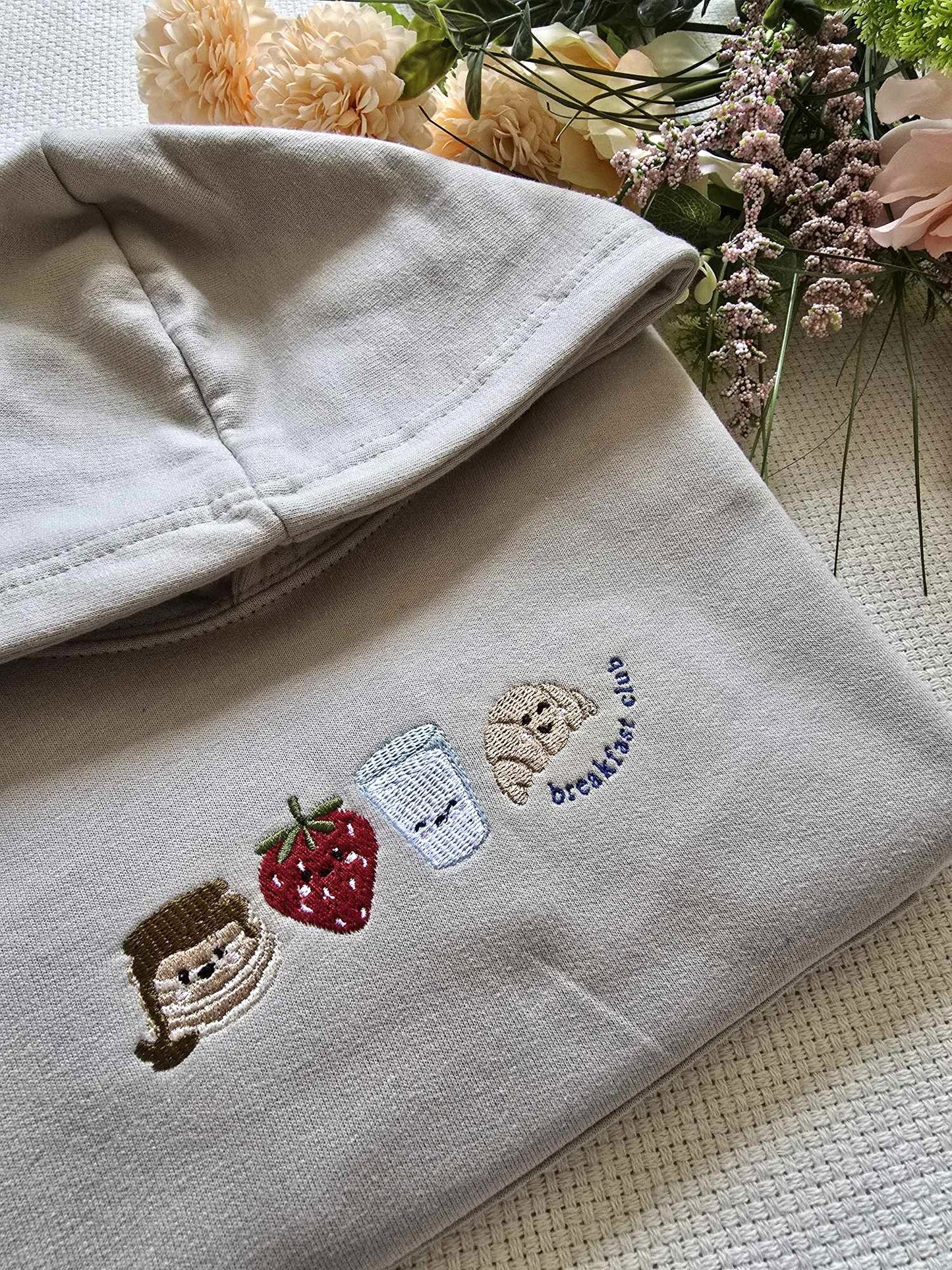 A beige sweatshirt with character and fruit embroidery, laid flat with flowers in the background.