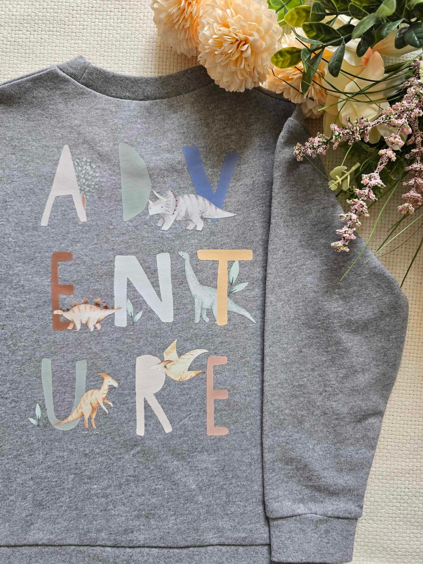 A grey sweatshirt with the word 'ADVENTURE' written on it, featuring dinosaur illustrations, displayed alongside some flowers.
