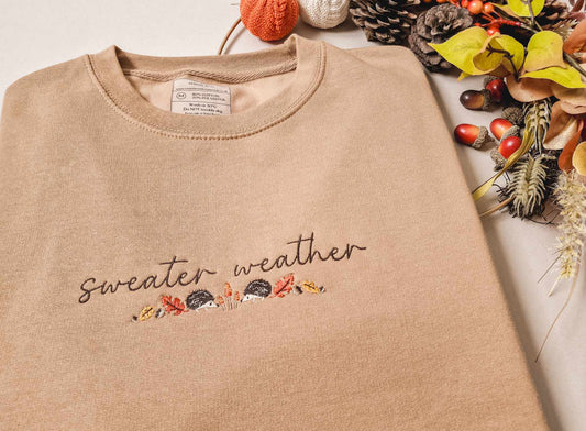 A beige embroidered sweatshirt with text 'SWEATER WEATHER' and autumn-themed decorations around it, displayed on a white surface with a floral arrangement and autumn leaves in the background.