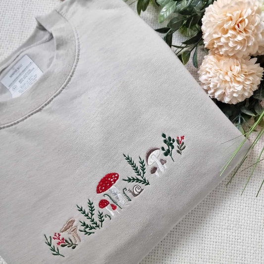 A beige sweatshirt with intricate embroidered designs featuring mushrooms, leaves, and other nature-themed elements, displayed alongside some flowers.