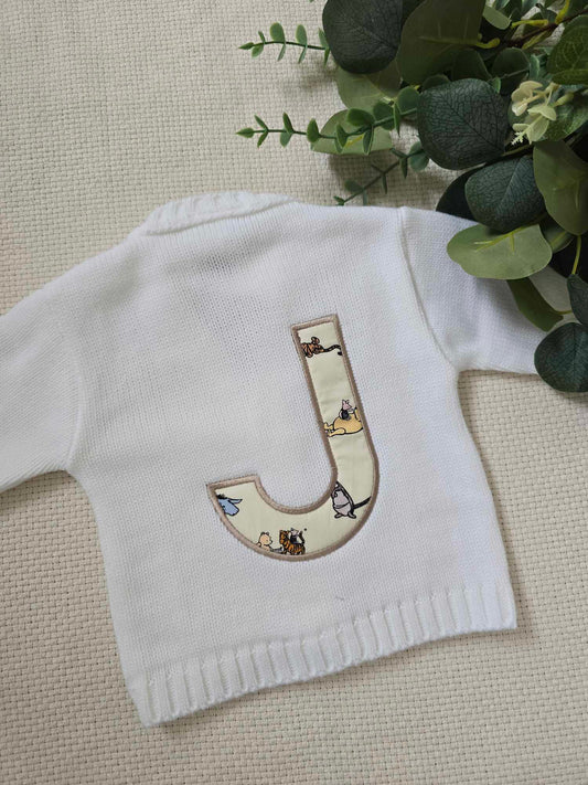 A white knitted cardigan with a Winnie the Pooh appliqué on the back, displayed on a flat surface with a green plant in the background.