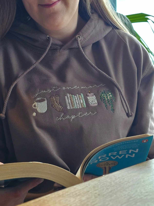 A person wearing a brown hoodie with embroidered designs including a cup of coffee and books, sitting at a table with an open book in front of them.