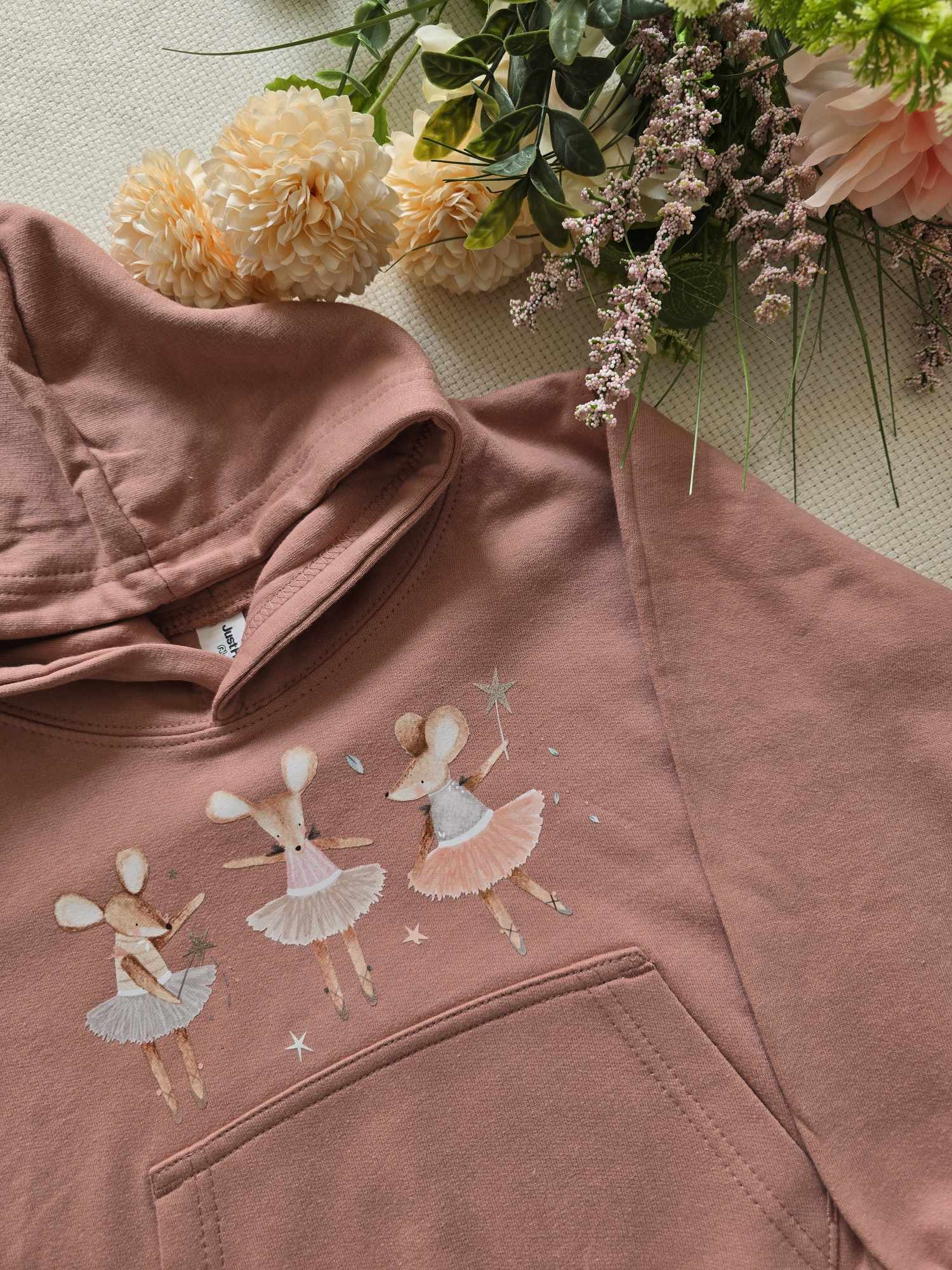 A pink sweatshirt with an embroidered design of ballet dancers and mice, presented alongside flowers.