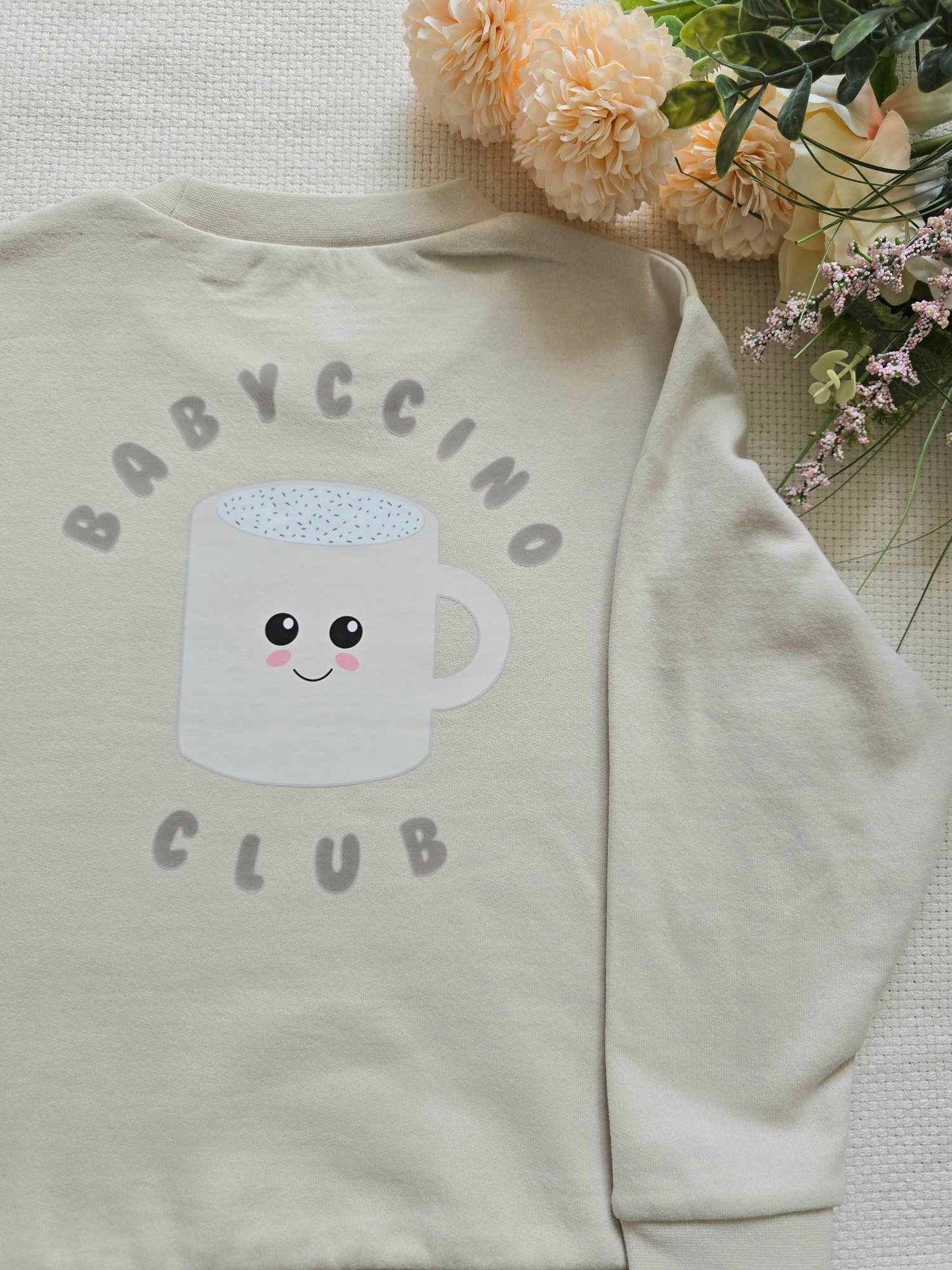 A beige sweatshirt with a graphic of a cup and the text 'BABYCCINO CLUB' on the front, displayed on a flat surface with flowers in the background.