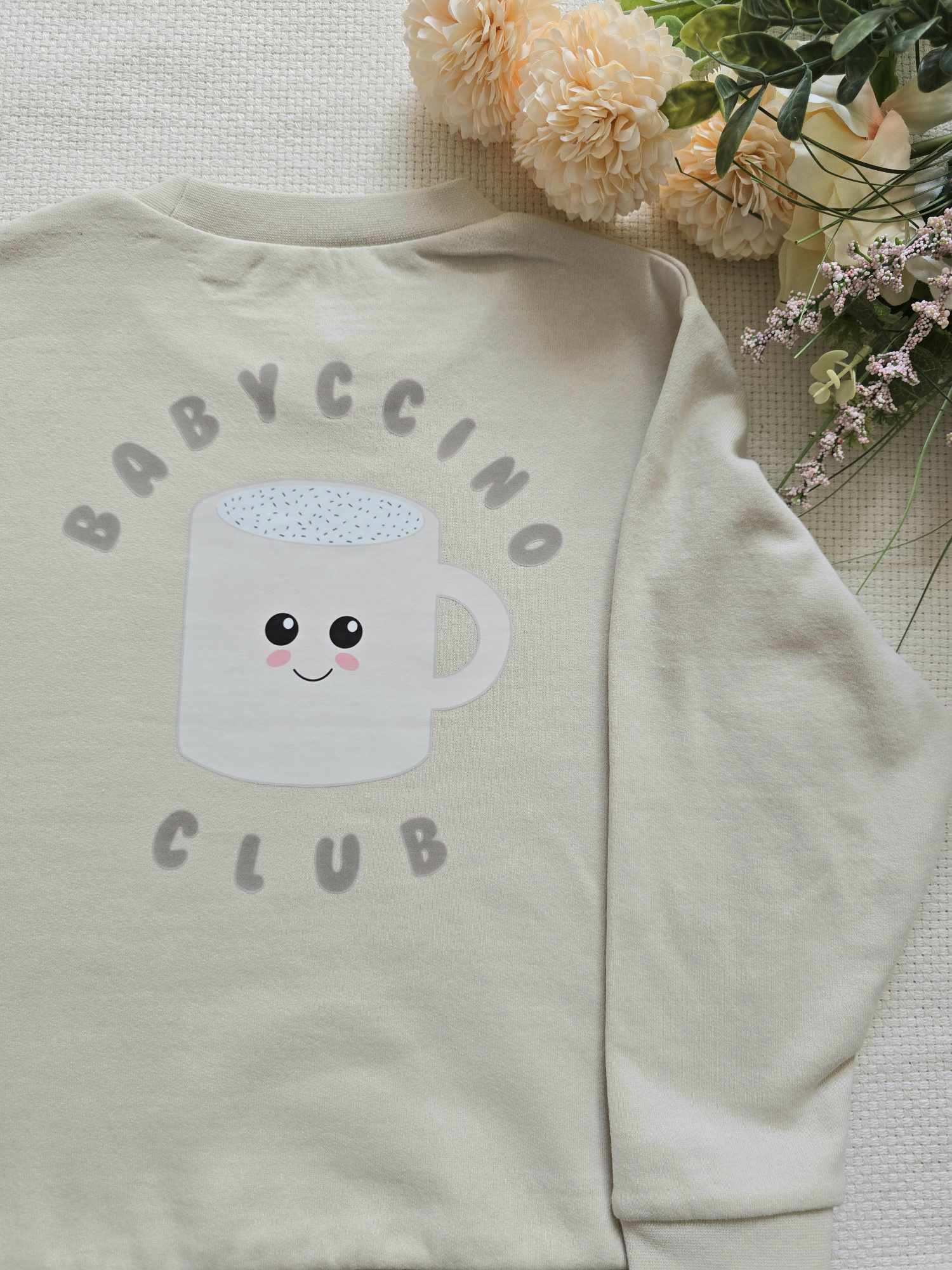 A beige sweatshirt with a graphic of a cup and the text 'BABYCCINO CLUB' on the front, displayed on a flat surface with flowers in the background.