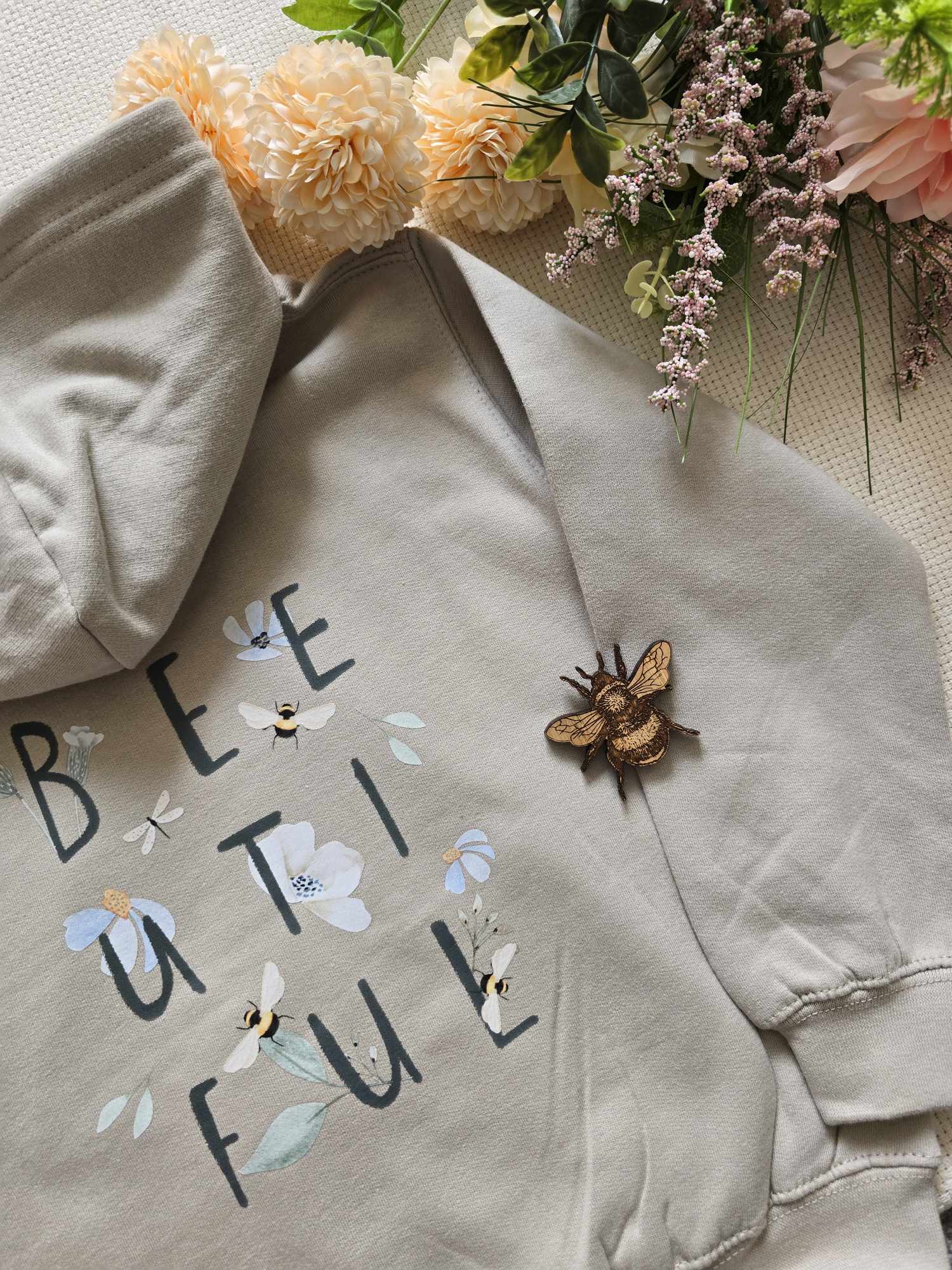 A beige sweatshirt with the text 'BEE UTLIFUL' on it, bees and flowers, laid flat with a floral arrangement in the background and a bee pin beside the text.