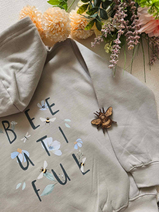 A beige sweatshirt with the text 'BEE UTLIFUL' on it, bees and flowers, laid flat with a floral arrangement in the background and a bee pin beside the text.
