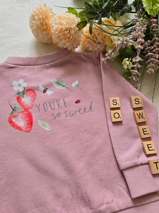 A pink sweatshirt with a text and strawberry graphic design, displayed alongside floral arrangements and strawberries.