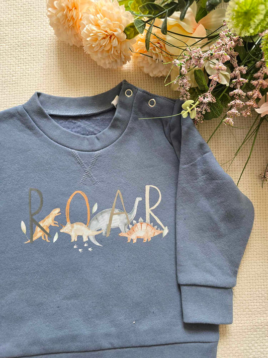A blue sweatshirt with the word 'ROAR' printed on the front with dinosaurs, laid flat with flowers in the background.