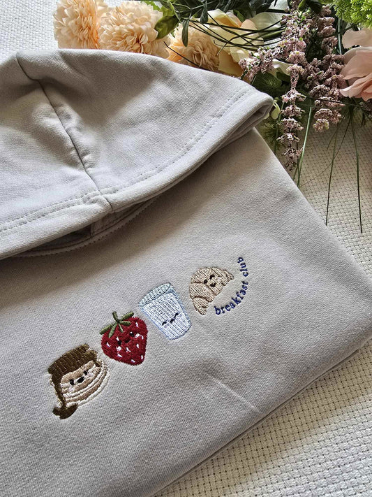 A beige sweatshirt with character and fruit embroidery, laid flat with flowers in the background.