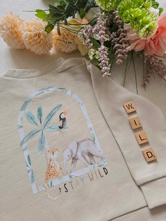 A beige sweatshirt with a graphic print of animals and the text 'STAY WILD', displayed alongside an elephant, cheetah toucan and palm tree.