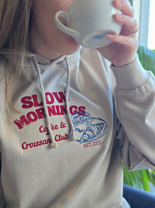A person wearing a beige hoodie with 'SLOW MORNINGS, COFFEE & CROISSANT CLUB' embroidered on the front, drinking from a mug.